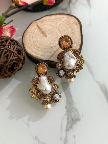 A glamorous set of earrings featuring irregular baroque pearls, large gemstone studs, and floral gold accents with crystal embellishments, displayed on a marble surface with wooden slices for contrast.