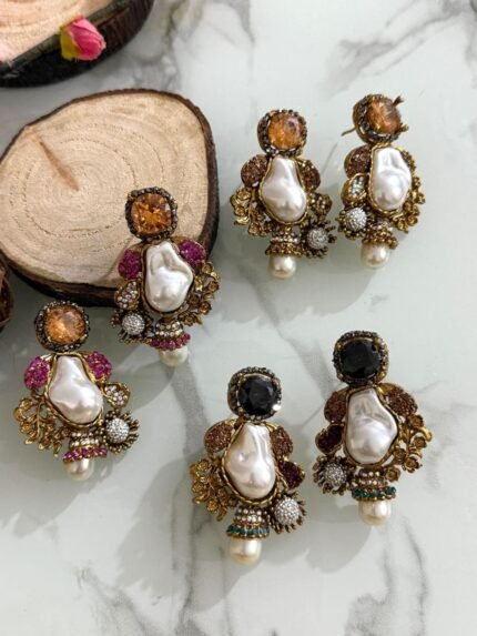 A glamorous set of earrings featuring irregular baroque pearls, large gemstone studs, and floral gold accents with crystal embellishments, displayed on a marble surface with wooden slices for contrast.