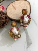 A glamorous set of earrings featuring irregular baroque pearls, large gemstone studs, and floral gold accents with crystal embellishments, displayed on a marble surface with wooden slices for contrast.