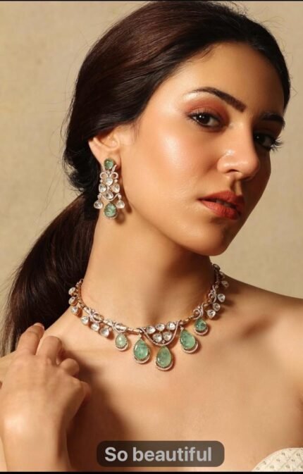 A traditional gold-tone kundan necklace with teardrop-shaped green emerald stones and shimmering white kundan detailing, worn with an ivory outfit.