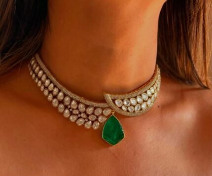 A crescent-shaped gold kundan choker adorned with white teardrop stones and a single green emerald pendant hanging in the center, worn on a bare neckline.