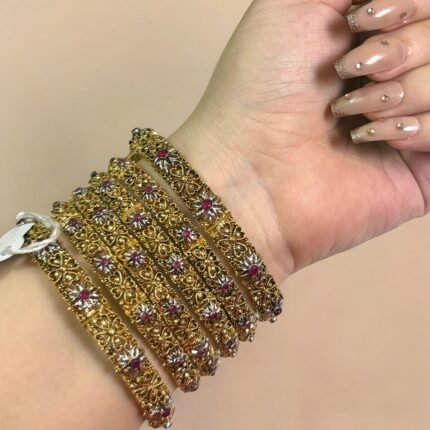 A hand adorned with six gold-plated bangles featuring intricate floral patterns and studded with red and silver stones, displayed against a light marble background.