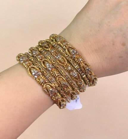 A hand wearing six intricately designed gold bangles featuring floral patterns adorned with small white stones. The bangles have a detailed leaf and petal motif throughout and are displayed against a white marble background. A tag with a price is tied to one bangle, and the nails are styled in nude polish with gold bead accents.