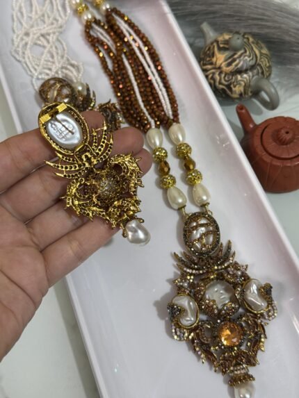 Three ornate antique-style necklaces with multistrand pearl and brown beads, featuring large gold-toned pendants embellished with pearls, gemstones, and intricate carved designs, displayed on a white tray.