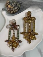 Pair of vintage-style dangler earrings featuring golden bird and floral designs, green stone inlays, pearls, and multicolor crystals, displayed on a white decorative shell plate.