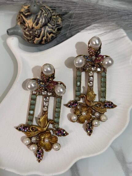 Pair of vintage-style dangler earrings featuring golden bird and floral designs, green stone inlays, pearls, and multicolor crystals, displayed on a white decorative shell plate.