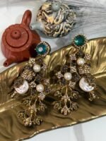 A pair of ornate, vintage-style drop earrings with emerald-green studs, crystal-studded floral motifs, and pearl accents, displayed on a golden leaf-shaped dish.
