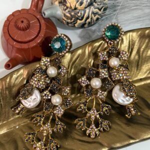 A pair of ornate, vintage-style drop earrings with emerald-green studs, crystal-studded floral motifs, and pearl accents, displayed on a golden leaf-shaped dish.
