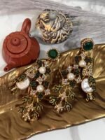 A pair of ornate, vintage-style drop earrings with emerald-green studs, crystal-studded floral motifs, and pearl accents, displayed on a golden leaf-shaped dish.