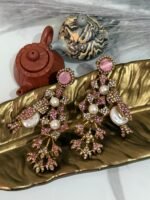 A pair of ornate, vintage-style drop earrings with emerald-green studs, crystal-studded floral motifs, and pearl accents, displayed on a golden leaf-shaped dish.