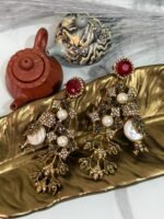 A pair of ornate, vintage-style drop earrings with emerald-green studs, crystal-studded floral motifs, and pearl accents, displayed on a golden leaf-shaped dish.