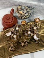 A pair of ornate, vintage-style drop earrings with emerald-green studs, crystal-studded floral motifs, and pearl accents, displayed on a golden leaf-shaped dish.