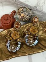 A pair of bold statement earrings with green gemstone studs, gold tiger heads, leaf accents, and circular bases decorated with iridescent shell inlay and floral designs.