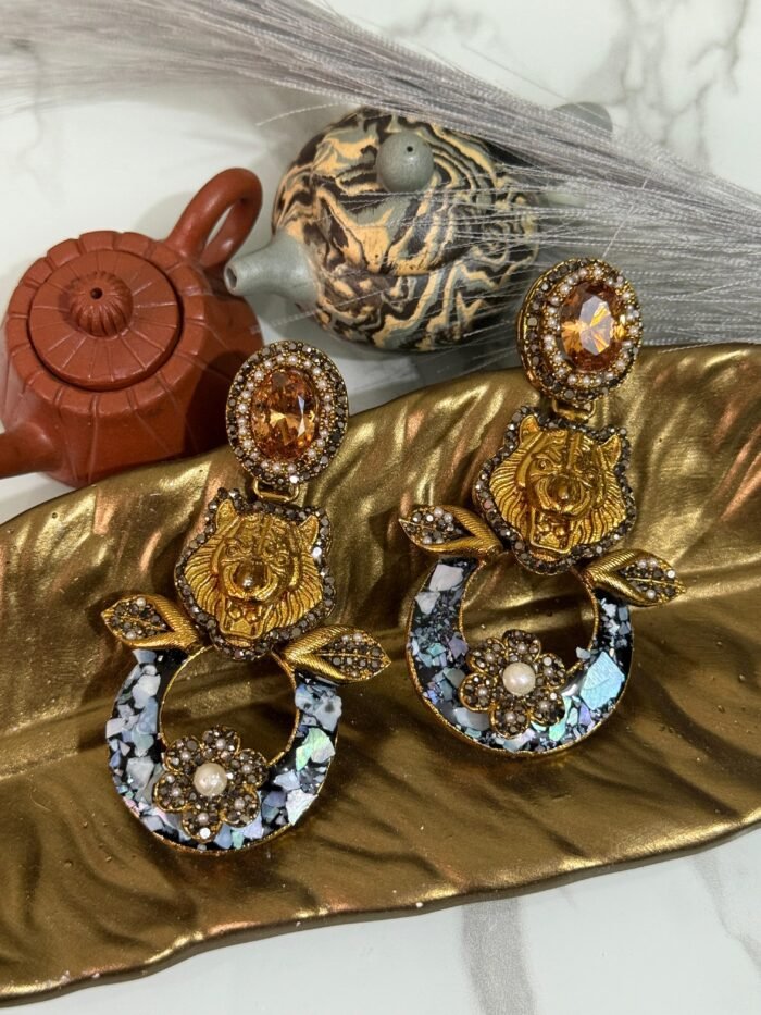 A pair of bold statement earrings with green gemstone studs, gold tiger heads, leaf accents, and circular bases decorated with iridescent shell inlay and floral designs.