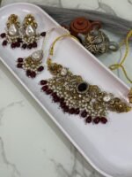 Traditional gold choker jewelry set with black center stone, maroon bead drops, pearls, and matching earrings and maang tikka, displayed in a white tray.