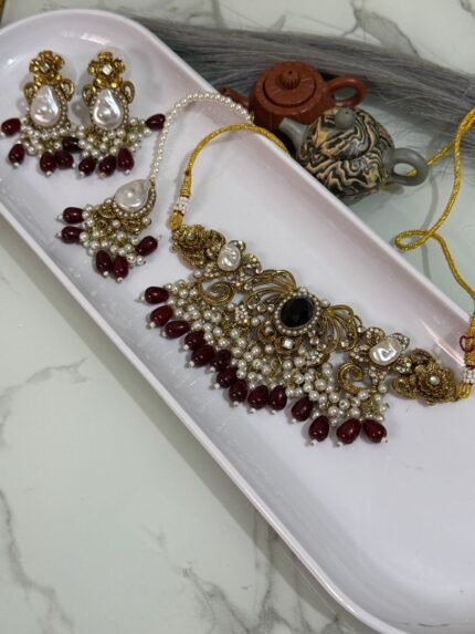 Traditional gold choker jewelry set with black center stone, maroon bead drops, pearls, and matching earrings and maang tikka, displayed in a white tray.