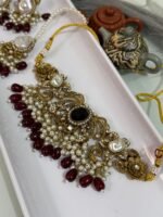 Traditional gold choker jewelry set with black center stone, maroon bead drops, pearls, and matching earrings and maang tikka, displayed in a white tray.