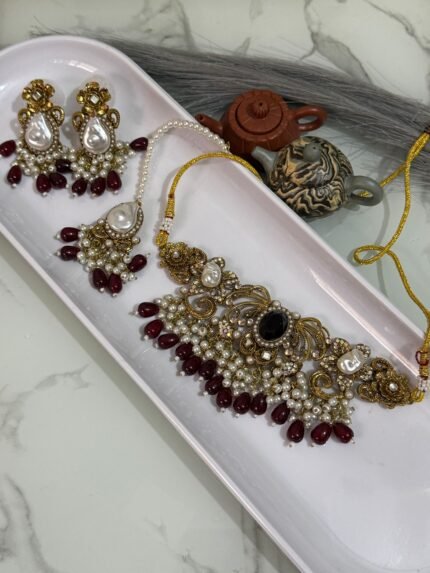 Traditional gold choker jewelry set with black center stone, maroon bead drops, pearls, and matching earrings and maang tikka, displayed in a white tray.