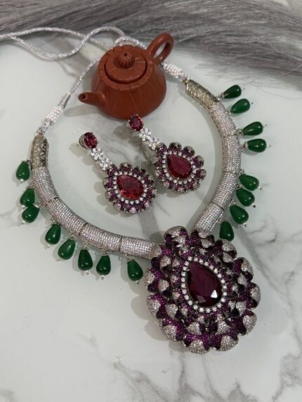 Ruby and diamond necklace set with green bead accents and matching earrings displayed on a marble surface.