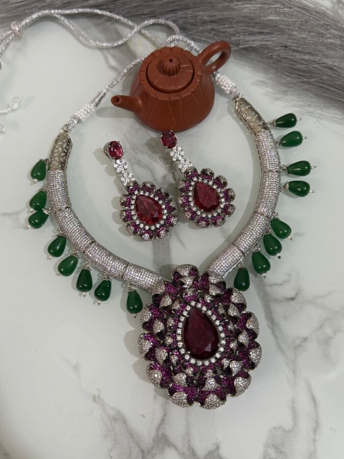 Ruby and diamond necklace set with green bead accents and matching earrings displayed on a marble surface.