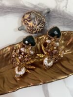 Gold-plated earrings with green stones, pearls, and floral design displayed on a golden leaf tray with decorative teapots in the background.