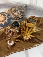 Gold-plated earrings with green stones, pearls, and floral design displayed on a golden leaf tray with decorative teapots in the background.
