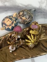 Gold-plated earrings with green stones, pearls, and floral design displayed on a golden leaf tray with decorative teapots in the background.