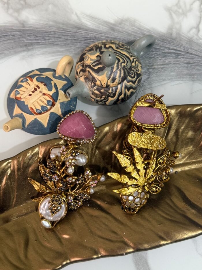 Gold-plated earrings with green stones, pearls, and floral design displayed on a golden leaf tray with decorative teapots in the background.
