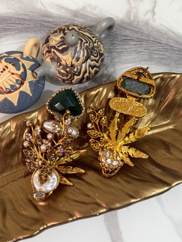 Gold-plated earrings with green stones, pearls, and floral design displayed on a golden leaf tray with decorative teapots in the background.