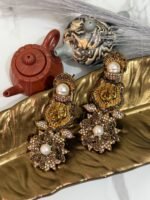 Gold earrings with a lion motif, pearl drop, and floral crystal design displayed on a golden leaf tray with teapots in the background.