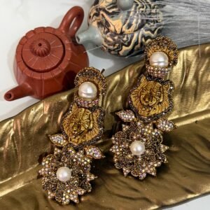 Gold earrings with a lion motif, pearl drop, and floral crystal design displayed on a golden leaf tray with teapots in the background.