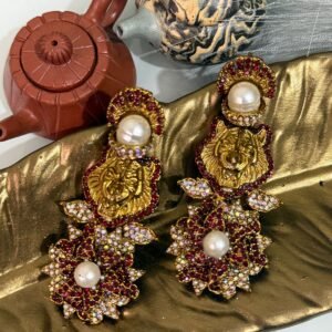 Gold earrings with a lion motif, pearl drop, and floral crystal design displayed on a golden leaf tray with teapots in the background.