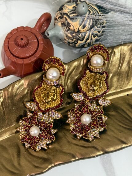 Gold earrings with a lion motif, pearl drop, and floral crystal design displayed on a golden leaf tray with teapots in the background.