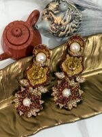 Gold earrings with a lion motif, pearl drop, and floral crystal design displayed on a golden leaf tray with teapots in the background.