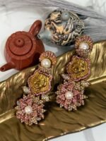 Gold earrings with a lion motif, pearl drop, and floral crystal design displayed on a golden leaf tray with teapots in the background.