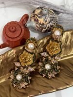 Gold earrings with a lion motif, pearl drop, and floral crystal design displayed on a golden leaf tray with teapots in the background.