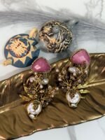 Gold-plated earrings with green stones, pearls, and floral design displayed on a golden leaf tray with decorative teapots in the background.