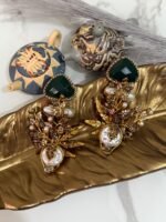 Gold-plated earrings with green stones, pearls, and floral design displayed on a golden leaf tray with decorative teapots in the background.