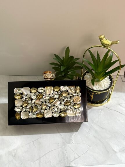 A luxurious jewel-encrusted clutch with a combination of crystal and pearlescent stones, set on a sleek black base, placed alongside decorative plants.