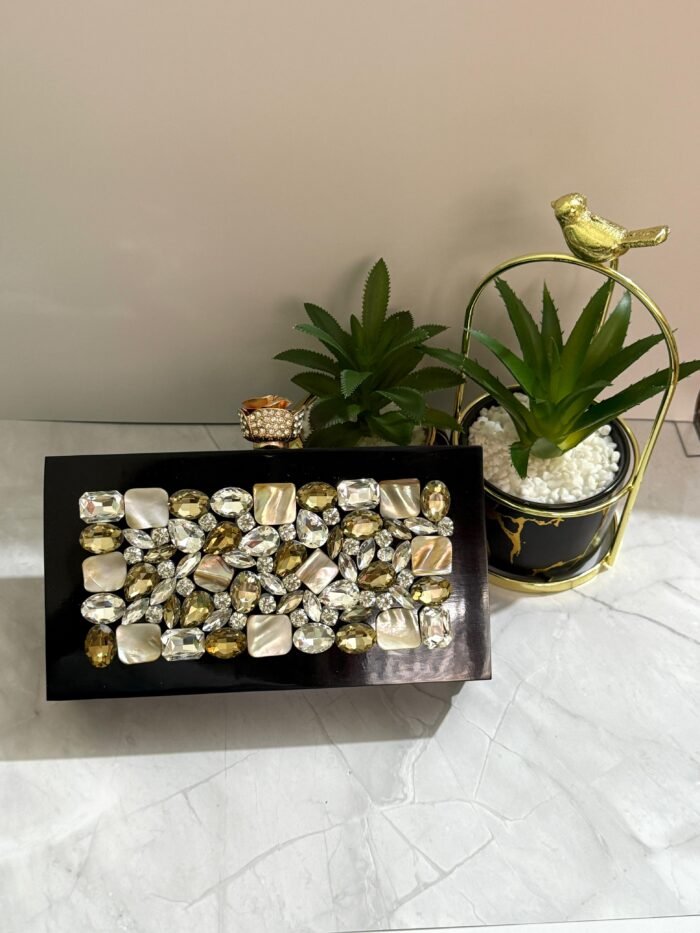 A luxurious jewel-encrusted clutch with a combination of crystal and pearlescent stones, set on a sleek black base, placed alongside decorative plants.
