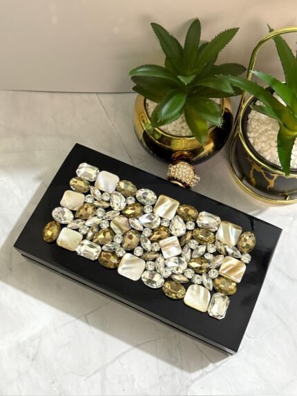 A luxurious jewel-encrusted clutch with a combination of crystal and pearlescent stones, set on a sleek black base, placed alongside decorative plants.