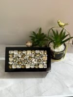 A luxurious jewel-encrusted clutch with a combination of crystal and pearlescent stones, set on a sleek black base, placed alongside decorative plants.