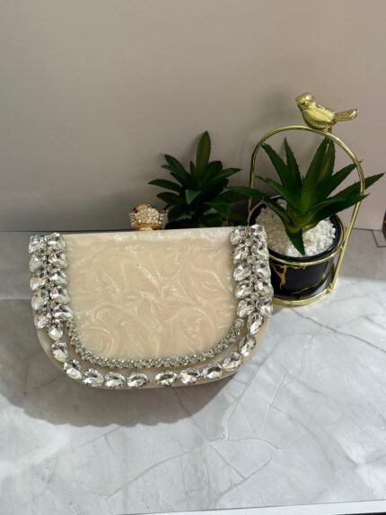 A luxurious cream-colored clutch with a row of sparkling clear crystals and pear-shaped gems, accompanied by decorative plants.