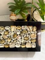 A luxurious jewel-encrusted clutch with a combination of crystal and pearlescent stones, set on a sleek black base, placed alongside decorative plants.