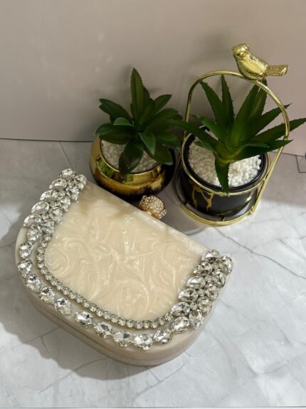 A luxurious cream-colored clutch with a row of sparkling clear crystals and pear-shaped gems, accompanied by decorative plants.