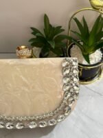 A luxurious cream-colored clutch with a row of sparkling clear crystals and pear-shaped gems, accompanied by decorative plants.