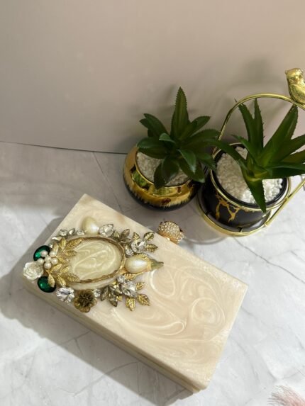 A vintage-inspired cream-colored clutch adorned with gold-tone leaves, pearls, and sparkling crystals, placed next to decorative plants.