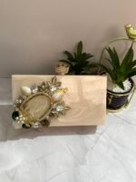 A vintage-inspired cream-colored clutch adorned with gold-tone leaves, pearls, and sparkling crystals, placed next to decorative plants.