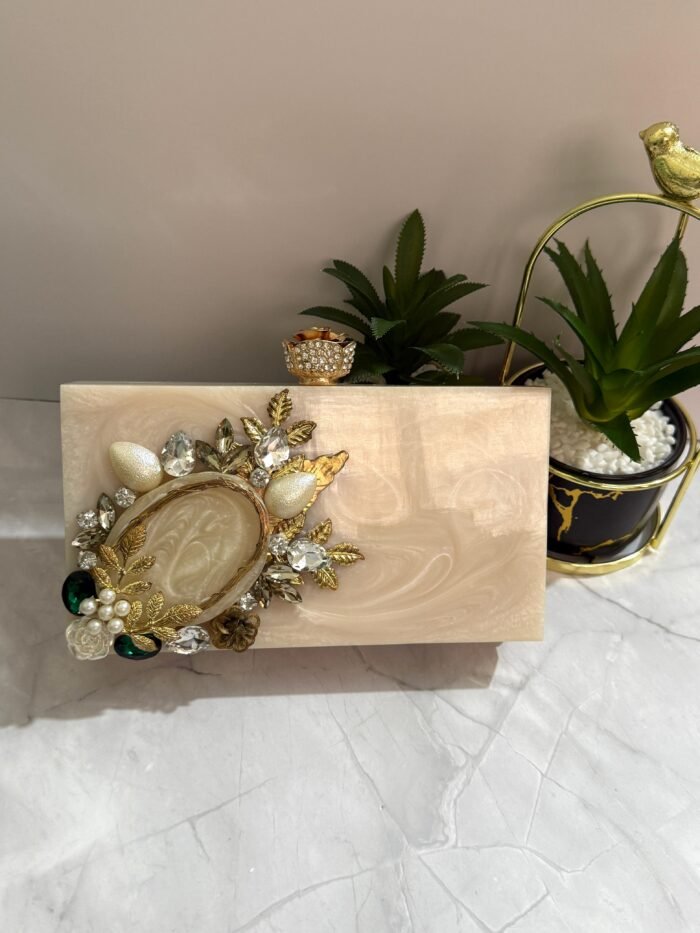 A vintage-inspired cream-colored clutch adorned with gold-tone leaves, pearls, and sparkling crystals, placed next to decorative plants.