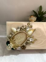 A vintage-inspired cream-colored clutch adorned with gold-tone leaves, pearls, and sparkling crystals, placed next to decorative plants.